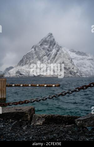 Stone embankment with chain fence and barrier against rippled water of strait washing coast with snowy mountain in winter overcast weather in Norway Stock Photo