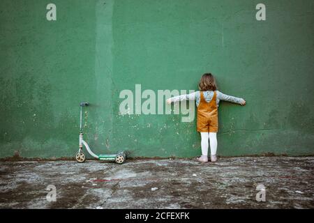 Back view of anonymous little girl with outstretched arms standing near weathered green wall and kick scooter and counting while playing hide and seek Stock Photo