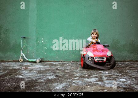 Cute little girl in casual clothes looking at camera and driving red electric vehicle with broken bumper against shabby green wall near kick scooter Stock Photo