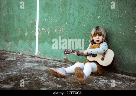 Little girl with ukulele sitting on rough ground near kick scooter against weathered green wall on street Stock Photo