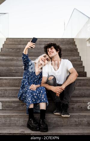 From below smiling trendy adult Woman and man in casual clothes taking selfie together with smartphone while sitting on wooden stairs Stock Photo
