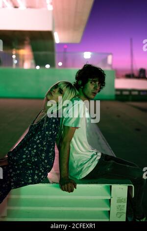 Attractive blond adult Woman in blue dress leaning on shoulder of handsome man in casual clothes while sitting and looking with curious gaze at camera against blurred purple sky and modern buildings during sunset Stock Photo