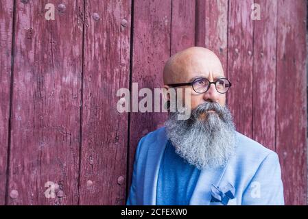 Front view of a mature trendy bearded hipster leaning on a vintage red door outdoors while looking away Stock Photo