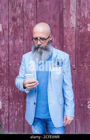 Front view of a mature trendy bearded hipster standing against a red vintage door outdoors while using a mobile phone Stock Photo