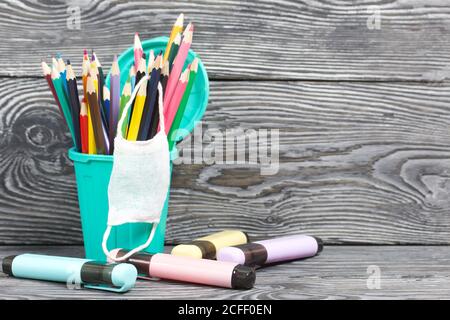 Colored pencils in a pencil holder. A medical mask is hanging on it. A pencil case in the form of a trash can. Nearby are colored markers. On brushed Stock Photo