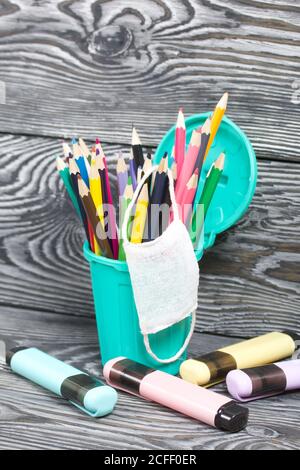 Colored pencils in a pencil holder. A medical mask is hanging on it. A pencil case in the form of a trash can. Nearby are colored markers. On brushed Stock Photo