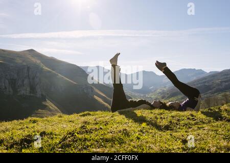 Side view of mother and daughter in sportswear doing exercise with raised legs while lying together on green grass of meadow in sunny mountains in Cantabria Stock Photo
