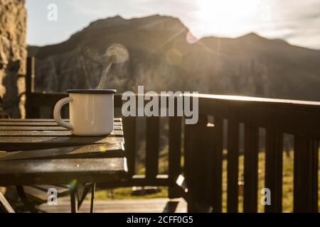 Steaming white mug with fresh hot beverage placed on wooden table on sunny terrace of stone house in sunny Cantabria Stock Photo