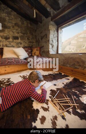 boy lying on floor on cozy carpet and drawing with colored pencils in sketchbook chilling cozy living room of stone house in Cantabria Stock Photo