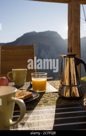 White cups and hot coffee maker placed on table with hot toast and glass of fresh orange juice on outdoor veranda with mountain on background Stock Photo