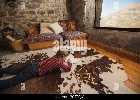 boy lying on floor on cozy carpet and drawing with colored pencils in sketchbook chilling cozy living room of stone house in Cantabria Stock Photo