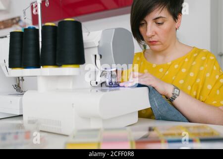 Focused brunette adult Woman smiling and using sewing machine to make denim garment while working in home workshop Stock Photo