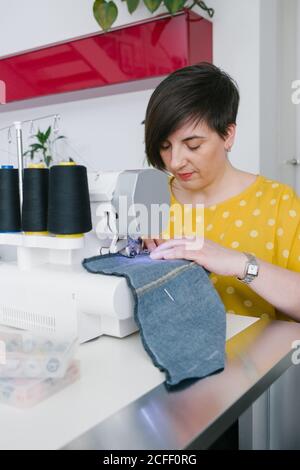 Focused brunette adult Woman smiling and using sewing machine to make denim garment while working in home workshop Stock Photo
