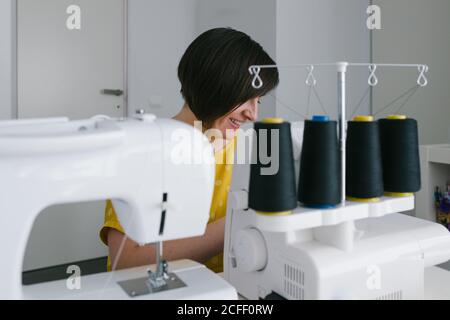Happy brunette adult Woman smiling and using sewing machine to make garment while working in home workshop Stock Photo