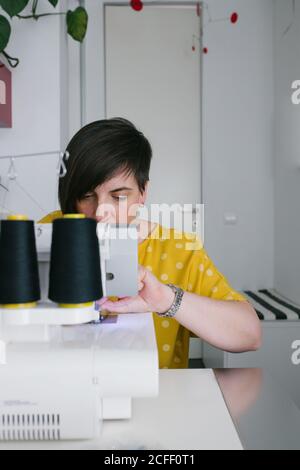 Focused brunette adult Woman smiling and using sewing machine to make garment while working in home workshop Stock Photo