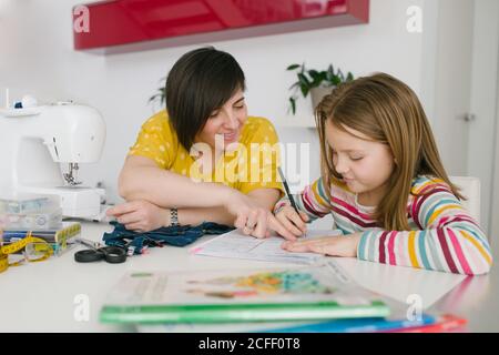 Happy adult Woman smiling and helping girl with homework assignment while sewing garment at home Stock Photo