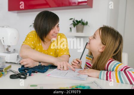 Happy adult Woman smiling and helping girl with homework assignment while sewing garment at home Stock Photo