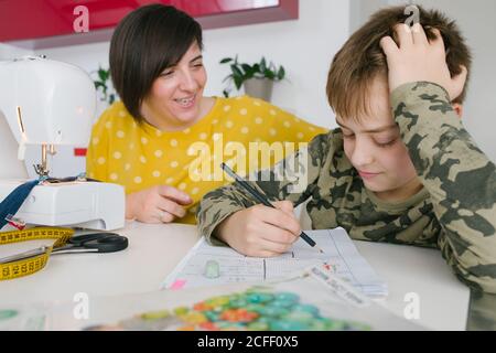 Focused boy doing homework assignment while sitting near adult Woman sewing garment at home Stock Photo