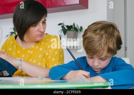 Focused boy doing homework assignment while sitting near adult Woman sewing garment at home Stock Photo