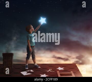 Fairy tale! The child hanging the stars in the sky. Boy on the roof cuts out stars. Stock Photo