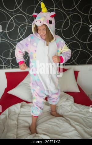 Happy joyful girl in funny kigurumi unicorn pajama playing in bedroom and jumping on bed with red and white bedding Stock Photo