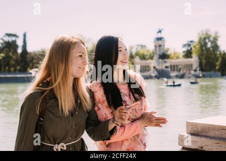 Multiethnic young women laughing and looking away while sitting on railing near river while spending time on embankment together Stock Photo