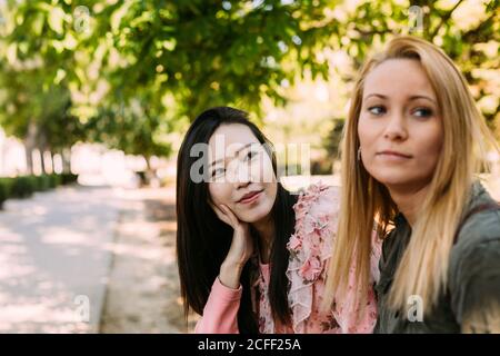 Two multiracial young women in casual outfits talking and looking at each other while sitting on bench in park Stock Photo
