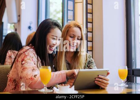 Young Caucasian Woman frowning and showing video on tablet to amazed Asian friend while sitting at cafe table together Stock Photo