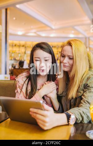 Young Caucasian Woman frowning and showing video on tablet to amazed Asian friend while sitting at cafe table together Stock Photo