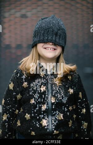 Funny anonymous kid in warm jacket and knitted cap covering half of face standing outdoors and smiling Stock Photo