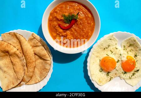 Famous traditional Arabic breakfast - peeled fava beans with chili pepper in white bowl, pita bread and fried eggs on a blue background. Top view Stock Photo