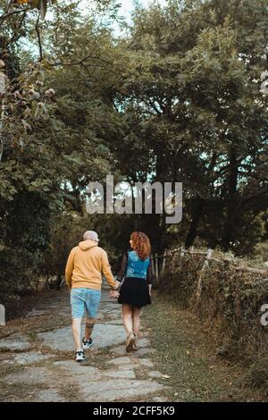 Back view of hipster couple spending happy time together while holding hands and walking on stone path among green trees Stock Photo
