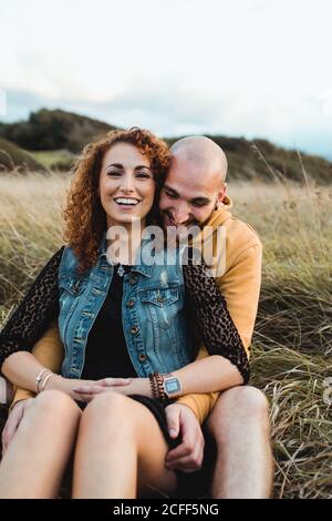 Young bald bearded man in yellow hoodie and jeans shorts embracing happy curly haired girlfriend in dress and denim vest while sitting together on meadow and talking Stock Photo