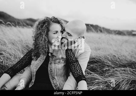 Young bald bearded man in yellow hoodie and jeans shorts embracing happy curly haired girlfriend in dress and denim vest while sitting together on meadow and talking Stock Photo