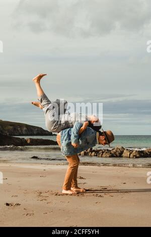 Side view of man holding on back laughing Woman with raised legs while having fun together and enjoying holidays on sandy beach with rocks on cloudy day in Scotland Stock Photo
