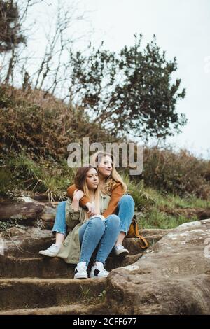 Relaxed women sitting on stone stairs on hill and hugging while resting after walking in countryside on cloudy spring day and enjoying time together on cloudy spring day Stock Photo