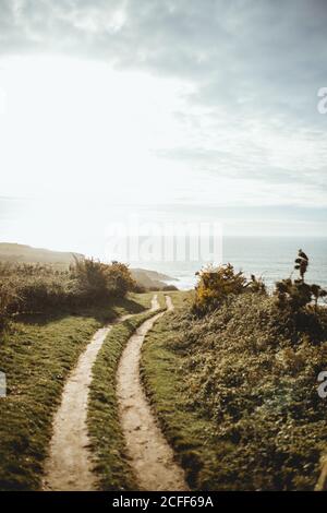 Empty rural dirt road with ruts leading through green spring field and hills to seaside at sunrise time with cloudy sky Stock Photo