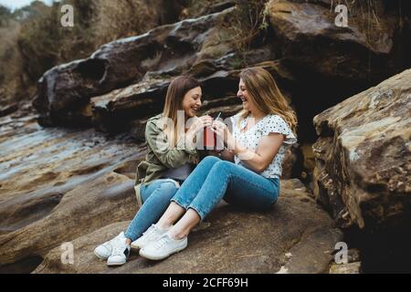 Relaxed women sitting on stone on hill sharing a beverage with straws while resting after walking in countryside on cloudy spring day and enjoying time together on cloudy spring day Stock Photo