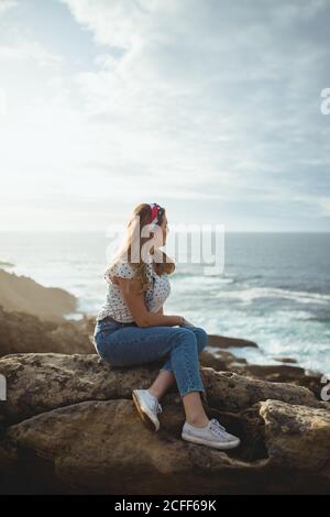 Side view of young female in casual clothes relaxing and dreaming while sitting on stone and looking at sea waves in sunny day with clouds on sky Stock Photo