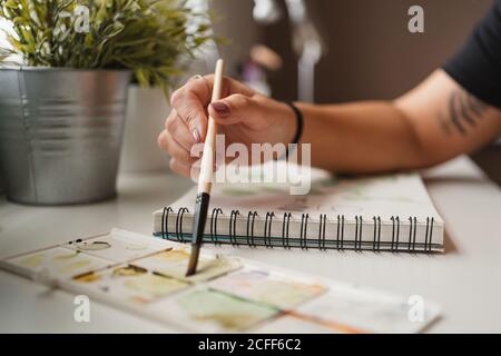 Closeup of unrecognizable woman using palette and watercolors while creating picture and sitting at table at home in Paris