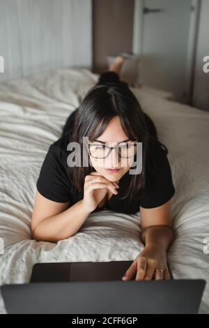 from above view of concentrated young Woman in glasses typing on laptop while studying and laying on bed at home in Paris