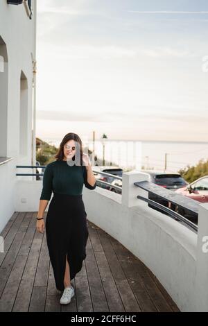 Thoughtful young female in casual look with black maxi skirt and white snickers walking on balcony with wooden floor while spending time in white stone house stayed at seaside or riverside Stock Photo