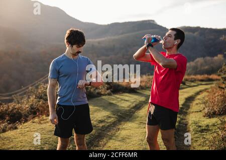 Adult athletic man standing on green trail and drinking water while friend look at the watch during training in mountains in sunny day Stock Photo
