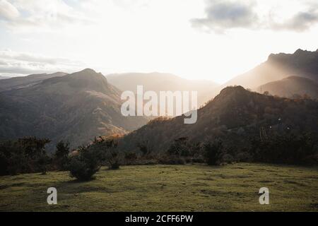 Scenic peaceful landscape with green valley and hills covered with forest and sun shining from behind mountains at early morning time with clouds on sky Stock Photo