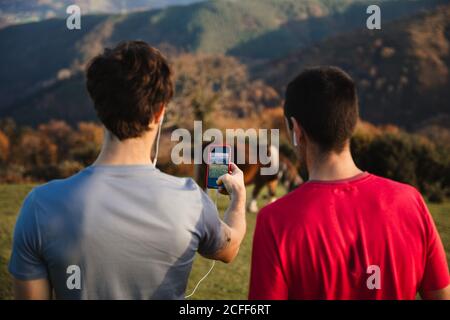Back view of sportive males standing on top of green hill taking picture with mobile phone of a cow on pasture while enjoying landscape relaxing after running in mountains Stock Photo