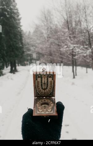 Crop hand of traveler in black knitted glove holding vintage compass with sundial while standing on snowy road among forest in gloomy winter day Stock Photo