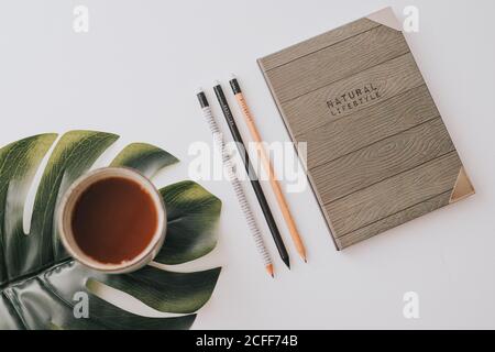 From above stylish notebook with wooden cover composed with row of pencils and mug of hot drink on green Monstera leaf on white background Stock Photo