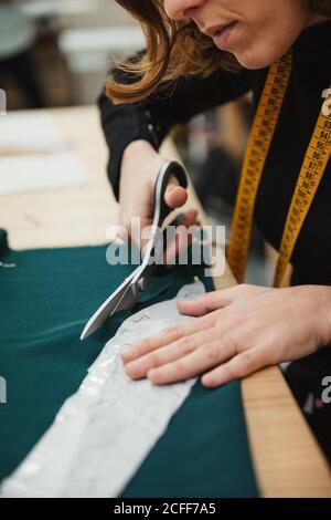 unrecognizable Woman using sharp scissors to cut garment detail from fabric while sitting at table in tailor workshop Stock Photo