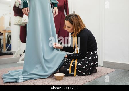 Dressmaker kneeling on carpet and fitting skirt of custom gown while working in professional studio Stock Photo