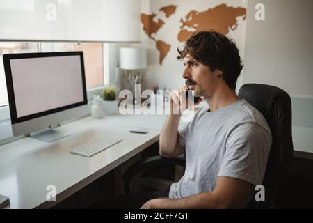 Side view of focused male freelancer sitting at table in creative workspace using mobile phone while working on online project Stock Photo